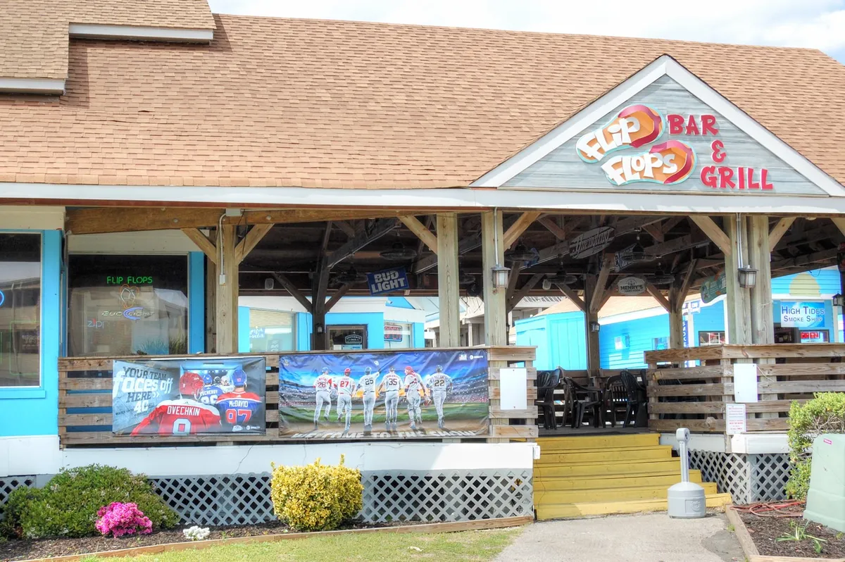 This image captures the exterior of a lively bar and grill, complete with a prominent sign, promotional banners, and an inviting outdoor seating area. The atmosphere appears vibrant, suggesting a place where guests can enjoy drinks, socialize, and listen to live music.