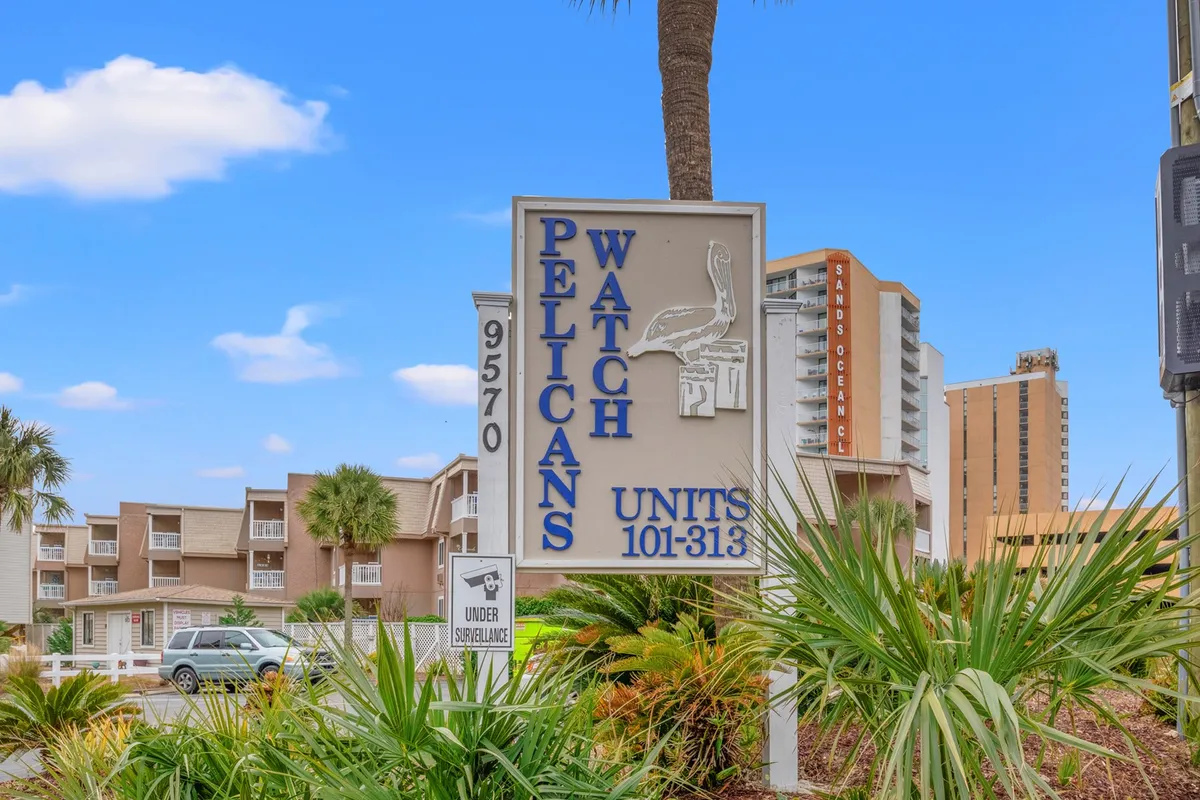 The welcoming entrance signage for Pelicans Watch is framed by lush palm trees and nearby buildings, setting the scene for a resort-like stay. The image hints at amenities like the pool and easy access to local attractions, while reinforcing the idea of a friendly, inviting complex.