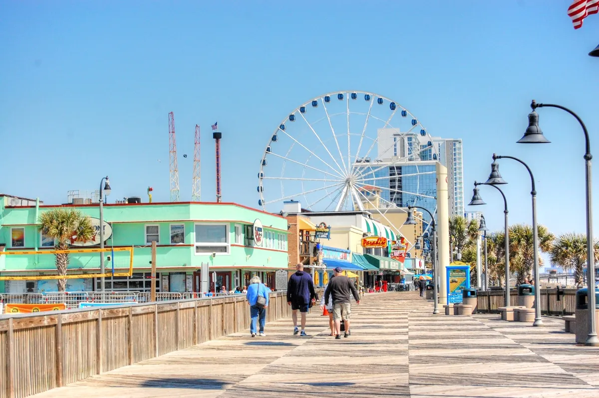 This image shows a bustling boardwalk scene, with a prominent ferris wheel, colorful buildings, palm trees, and visitors enjoying amusement park rides and the lively atmosphere.