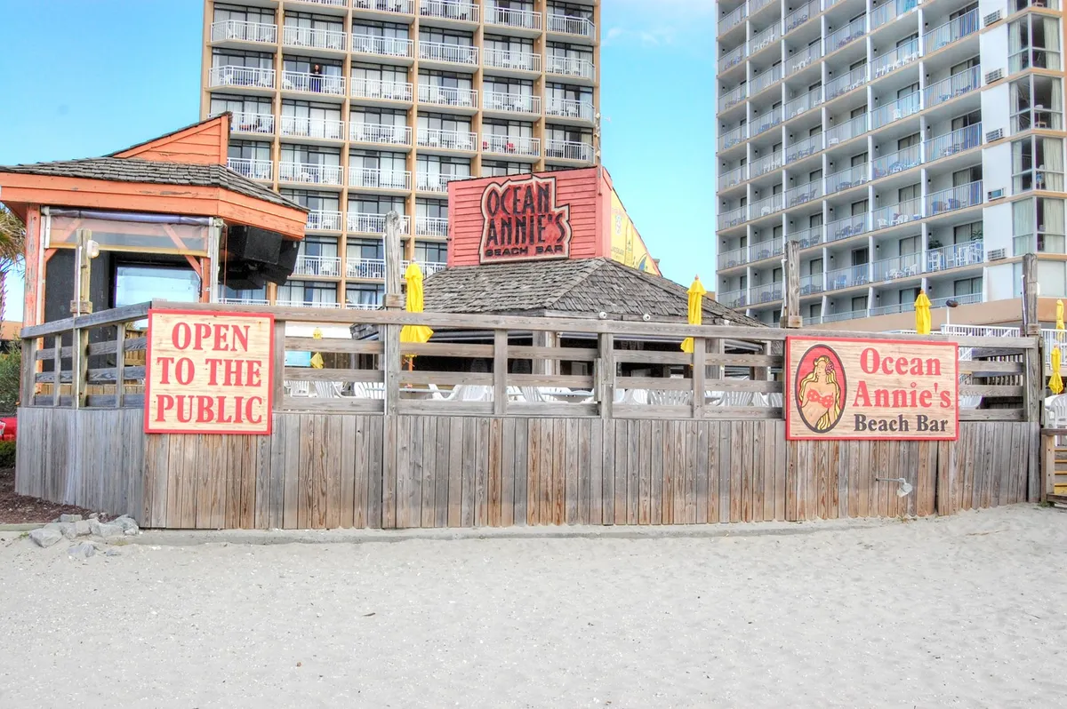 An exterior scene featuring the recognizable Ocean Annie's Beach Bar sign with wooden fencing, an outdoor seating area, and cheerful yellow umbrellas. The setting invites guests to enjoy live music, casual dining, and direct access to the beach, all in a relaxed, welcoming atmosphere.