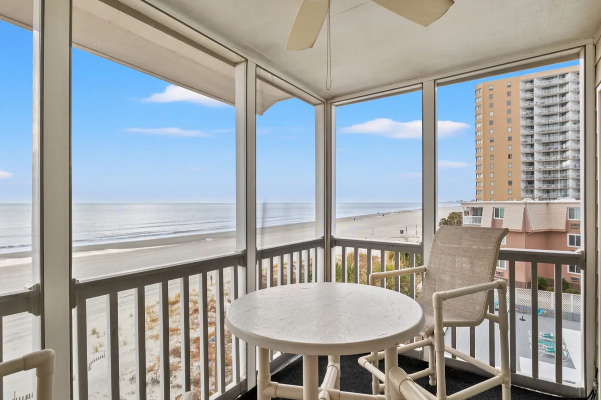 This image captures a serene screened-in balcony furnished with a table and chairs, ceiling fan overhead, and open views of the coastline, sandy beach, and waves beyond. The vantage point demonstrates the seamless transition from the comfort of the balcony to the vibrant ocean scene outside.