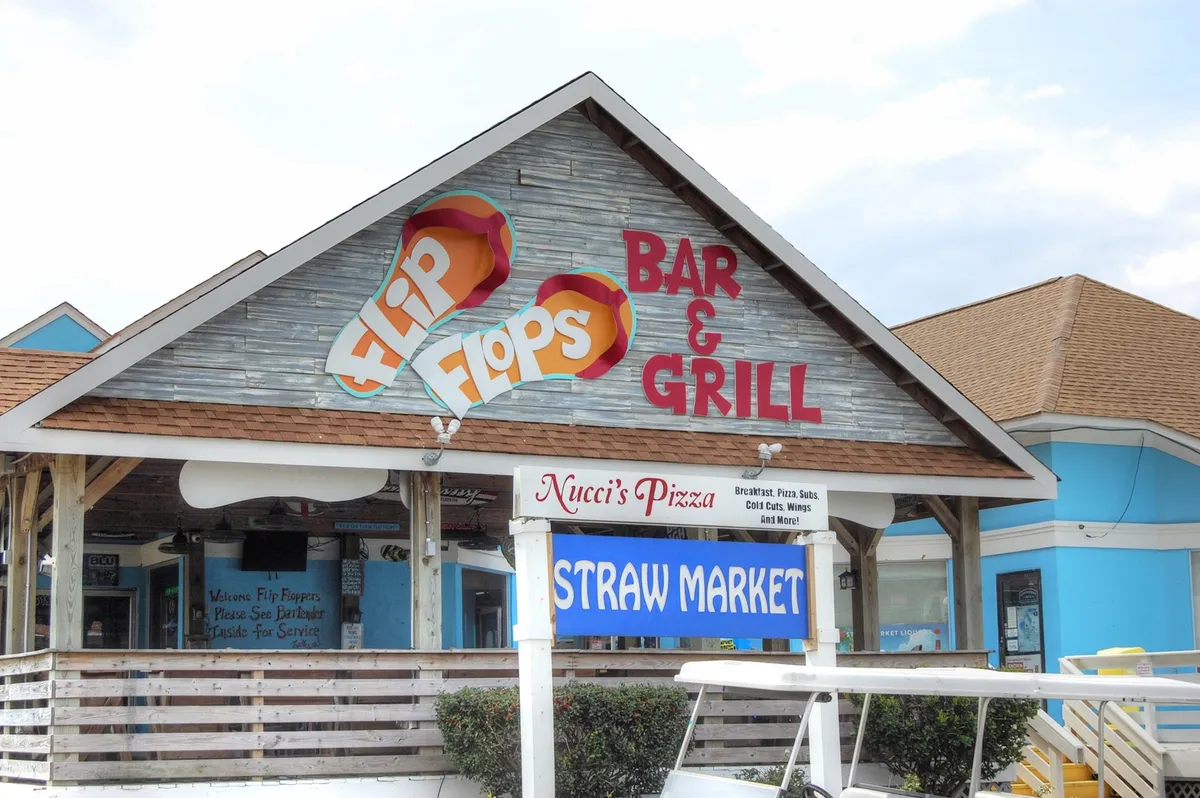 A lively exterior view showcasing the signs for Flip Flops Bar & Grill, Nucci's Pizza, and the Straw Market. The scene captures the vibrant local dining and shopping options, with people gathering and enjoying relaxed, communal spaces.