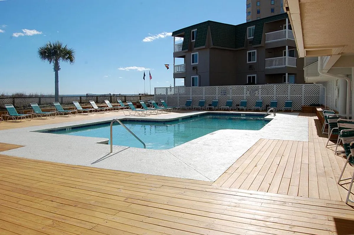 A vibrant outdoor swimming pool area surrounded by palm trees with plenty of lounge chairs for sunbathing. The pool is flanked by the resort’s buildings, reflecting a lively, inviting atmosphere ideal for both relaxation and socializing.