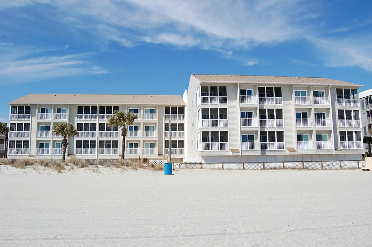 This exterior shot captures beachfront condominium buildings set against a backdrop of palm trees, soft sand, and a clear blue sky. The inviting shoreline is just steps away, emphasizing ease of beach access and the relaxing vacation atmosphere. Families can be imagined lounging on the sand, playing games, or heading back to the buildings after a fun beach day.