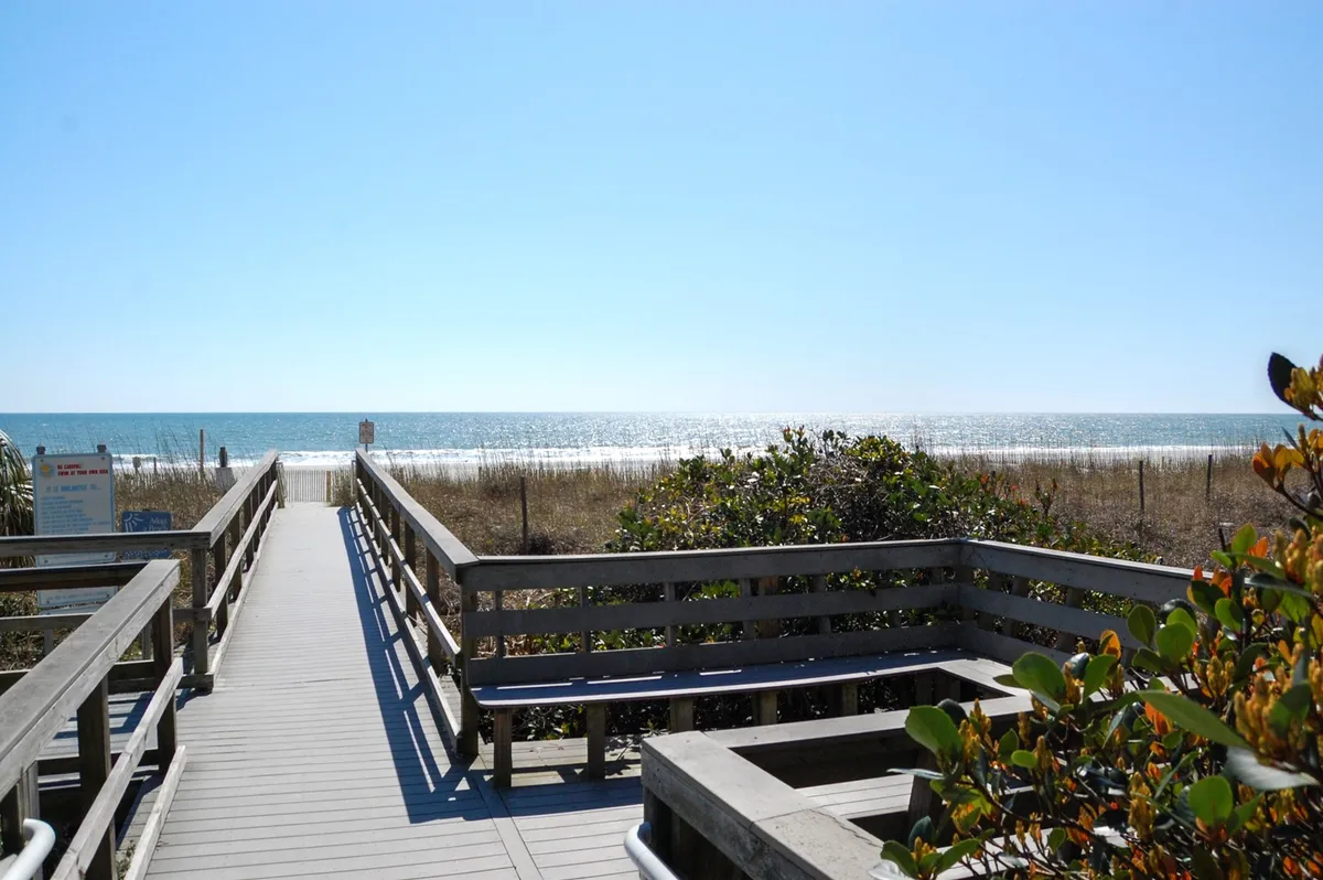 A wooden boardwalk leading through dunes and vegetation toward the ocean, with expansive views of the beach and water beyond. The scene is calm and inviting, capturing the anticipation and beauty of direct beach access.