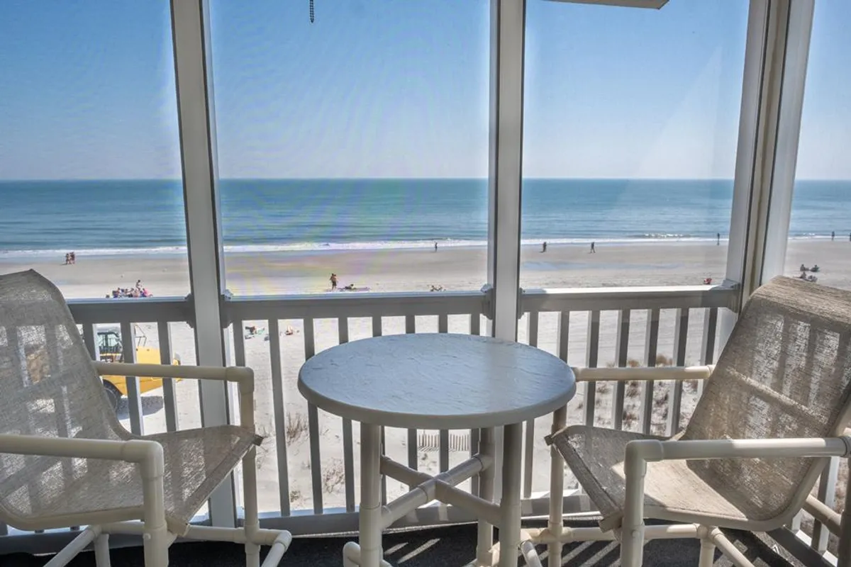 A cozy screened-in balcony with two rocking chairs and a circular table, offering unbroken vistas of the ocean and sky. The scene is peaceful, ideal for relaxing with a book or enjoying a quiet conversation while taking in the sea breeze.