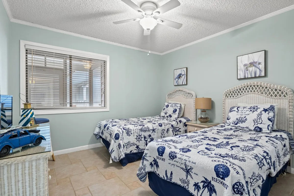 A cheerful bedroom with two twin beds adorned in ocean-themed bedding, wicker headboards, a softly glowing bedside lamp, framed artwork, and a vintage toy car on a side table. The room is bright, inviting, and perfect for families or children.