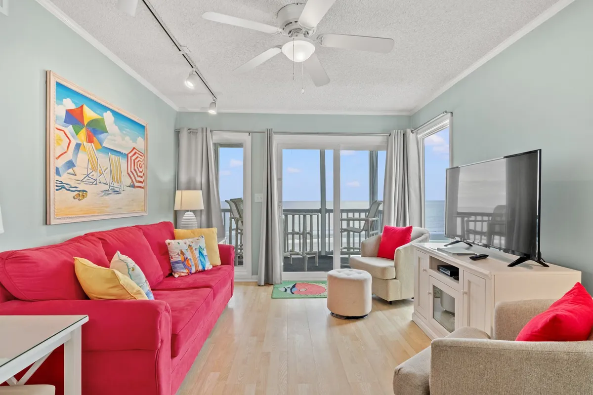 The image showcases a sunlit living room with a striking red sofa facing large windows that offer sweeping ocean views. Cheerful, colorful artwork adorns the walls, and there's a television for entertainment. The atmosphere is bright and inviting, with plenty of natural light streaming in, creating the perfect spot to relax, enjoy the view, or gather for a cozy evening.