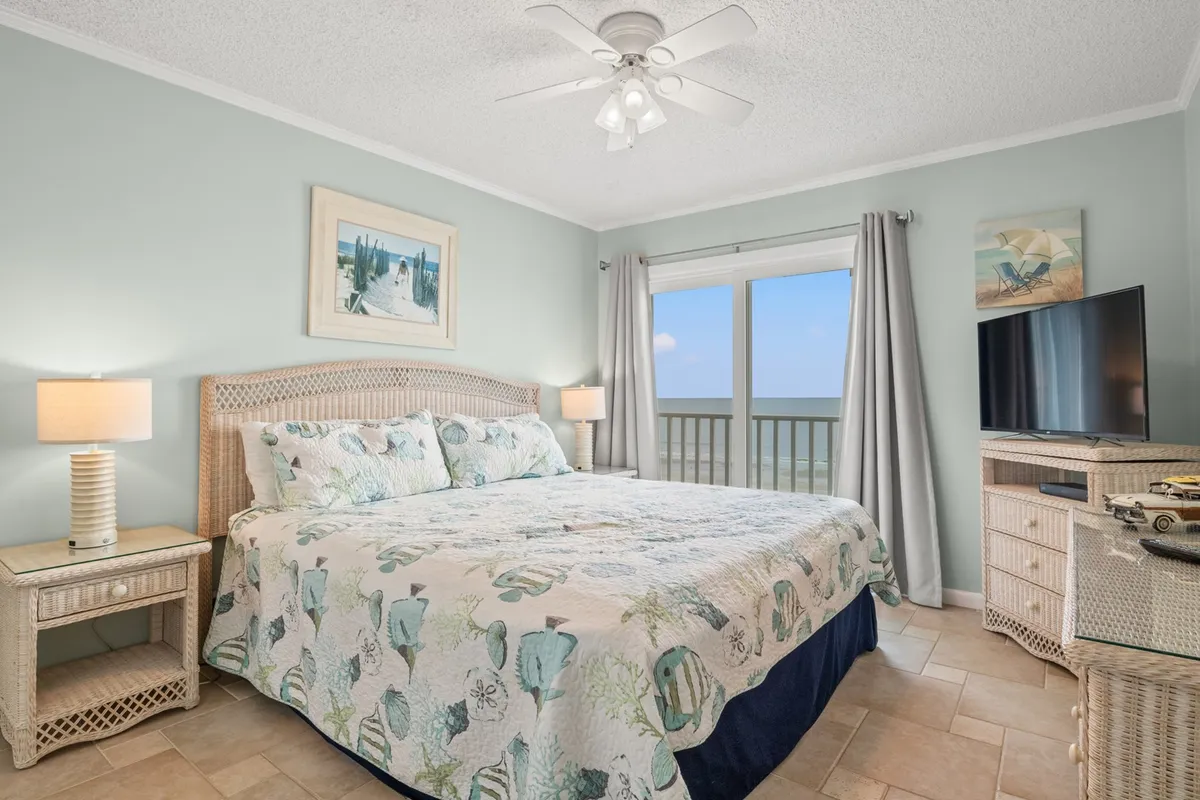 A serene bedroom featuring a bed with a sea-themed quilt, a large window that frames an ocean view, a nightstand equipped with a lamp, and a TV console. The natural light fills the space, enhancing the tranquil atmosphere ideal for rest and relaxation.