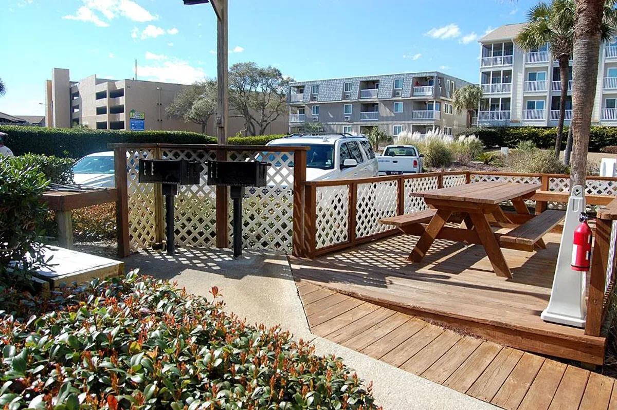 A landscaped outdoor area featuring picnic tables and built-in grilling stations, perfect for barbecuing or gathering with friends and family. Greenery surrounds the area, adding to the casual and friendly vibe.