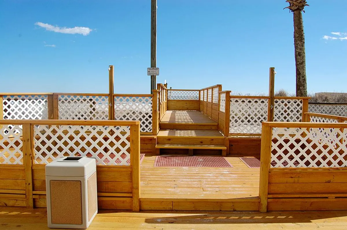 A cheerful exterior view of a wooden deck with lattice railings, palm trees in the background, and an inviting area for relaxing or socializing outdoors. The setup suggests a perfect backdrop for group conversations, morning coffee, or evening get-togethers.