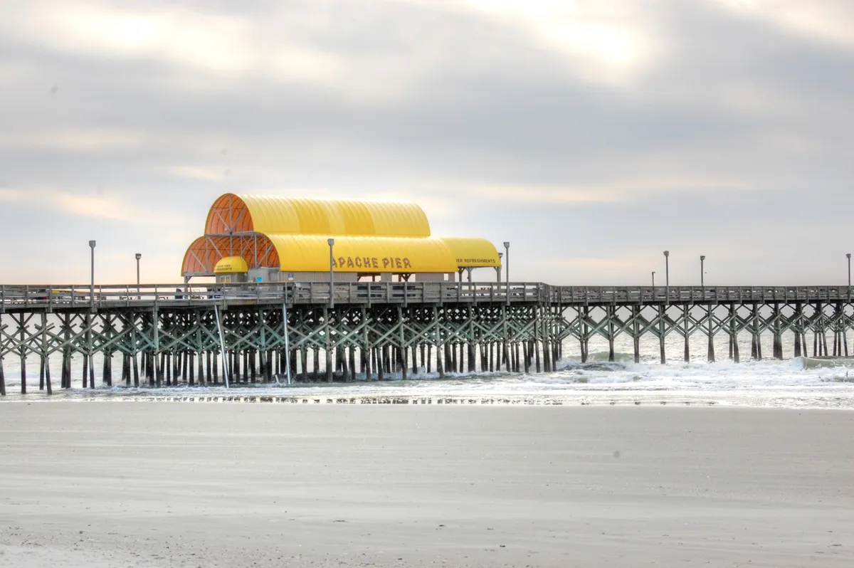 The photo captures Apache Pier with its distinctive yellow roof structure stretching over ocean waves. The pier is busy with people fishing, walking, and taking photographs, set against a relaxed beach backdrop.