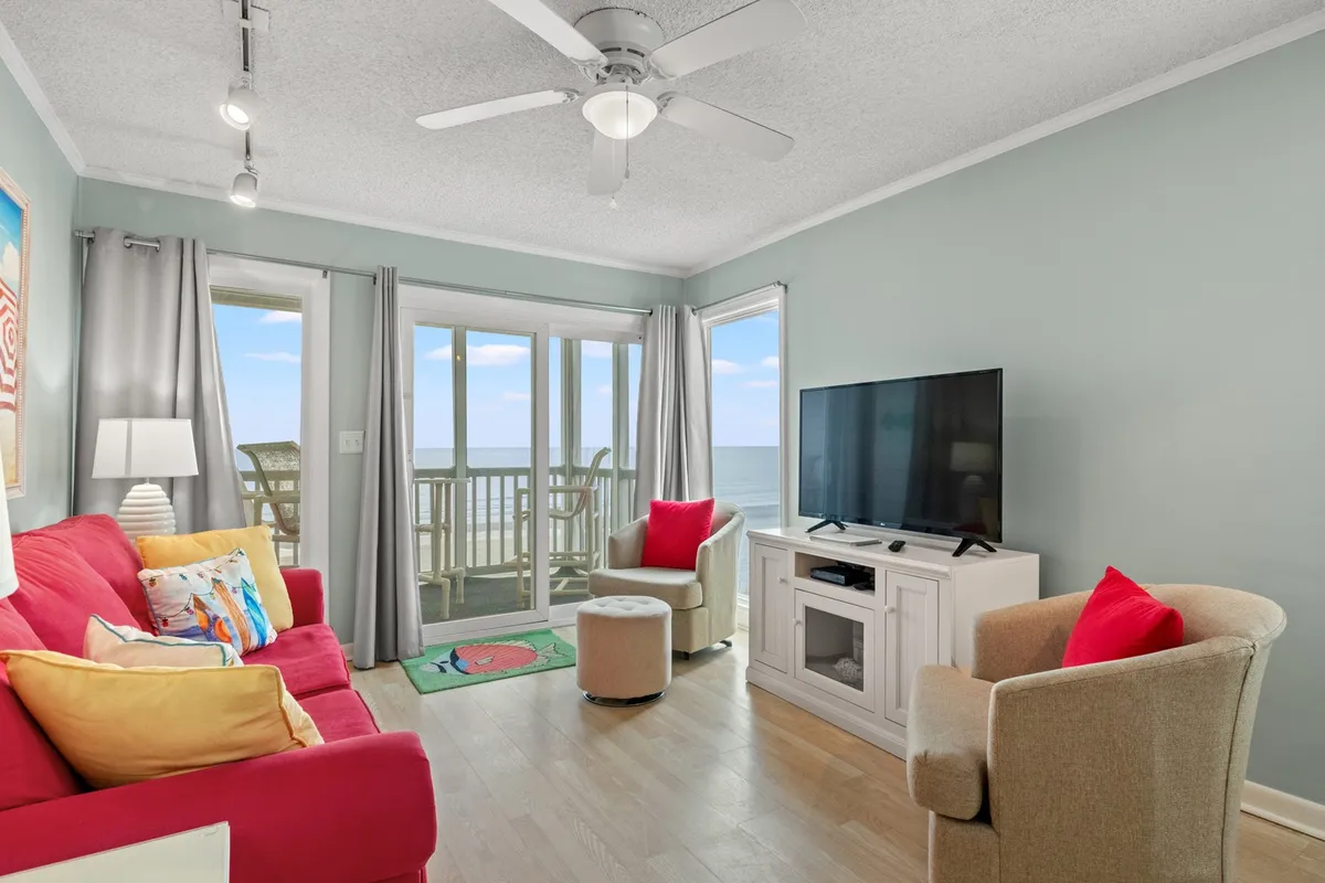 The image showcases a cheerful, open-plan living room with a bold red sofa positioned to take in panoramic ocean views through large windows. Decorative cushions invite relaxation, while a television is ready for movie nights or casual entertainment. The space is bathed in natural light, creating a warm and welcoming atmosphere that enhances the feeling of comfort and togetherness.