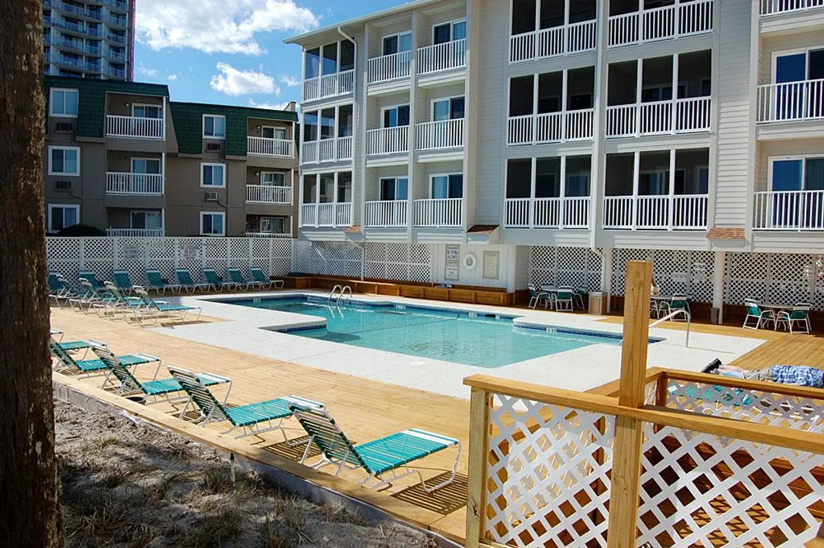 A wide view of the building's exterior shows balconies overlooking a sparkling swimming pool, with lounge chairs positioned around the pool deck. The scene illustrates how the balcony space is oriented in relation to the pool and emphasizes the inviting atmosphere below.
