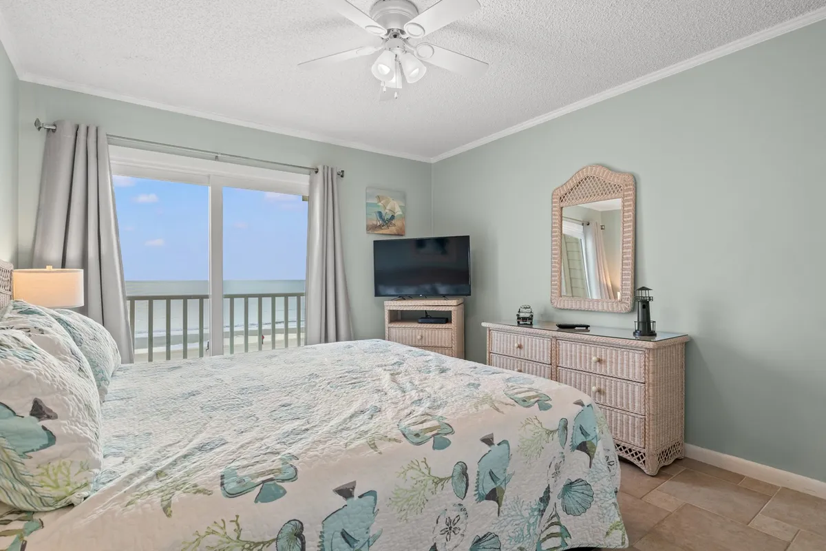 A tranquil bedroom with floral bedding, a large window offering sweeping ocean views, and a dresser with a mirror. The space is bright, serene, and oriented toward enjoying the natural beauty just outside the window.